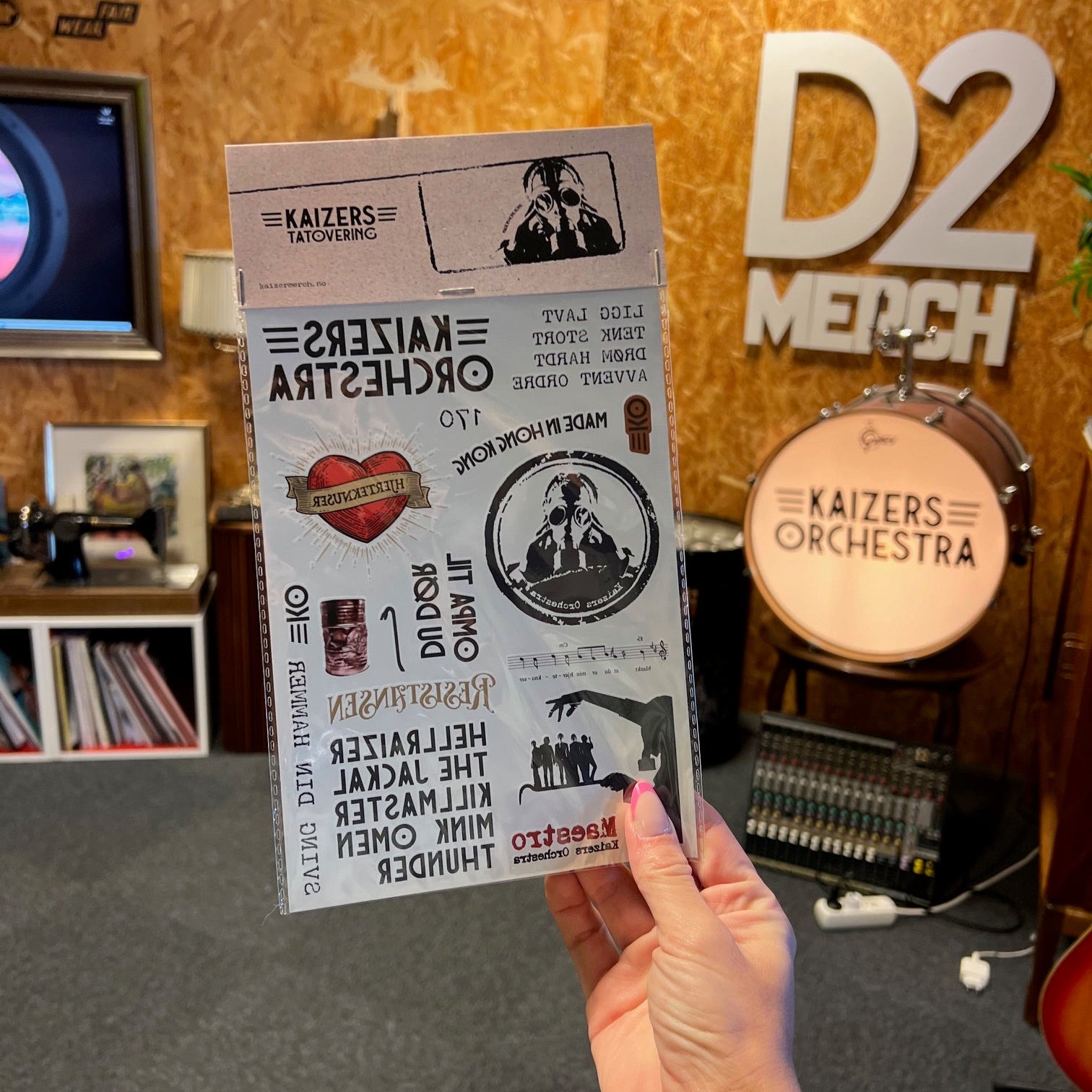 Person holding a set of temporary tattoos for The Kaizers Orchestra in front of a wooden counter with band name branding.
